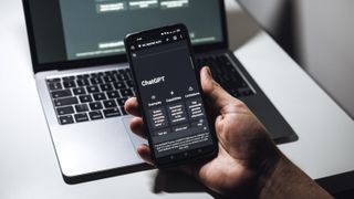 A hand holding a phone running ChatGPT in front of a laptop