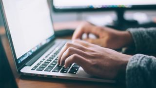 A laptop viewed from the side with a person typing on it. Their hands are lit up by the laptop's backlight