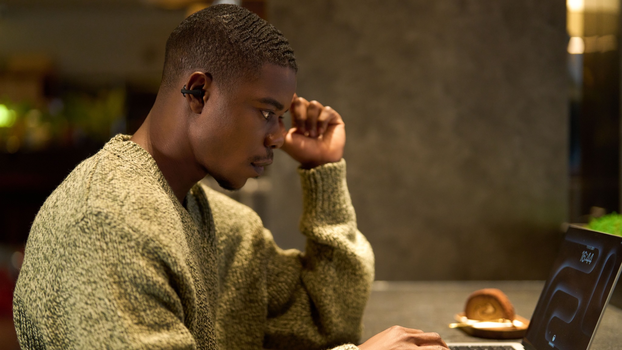 A young man with very short black hair wearing an oatmeal colored sweater is sat at a desk, staring intently at an Apple laptop while wearing a Shokz OpenDots ONE earbud. He must be very bored because the laptop is showing the lock screen.