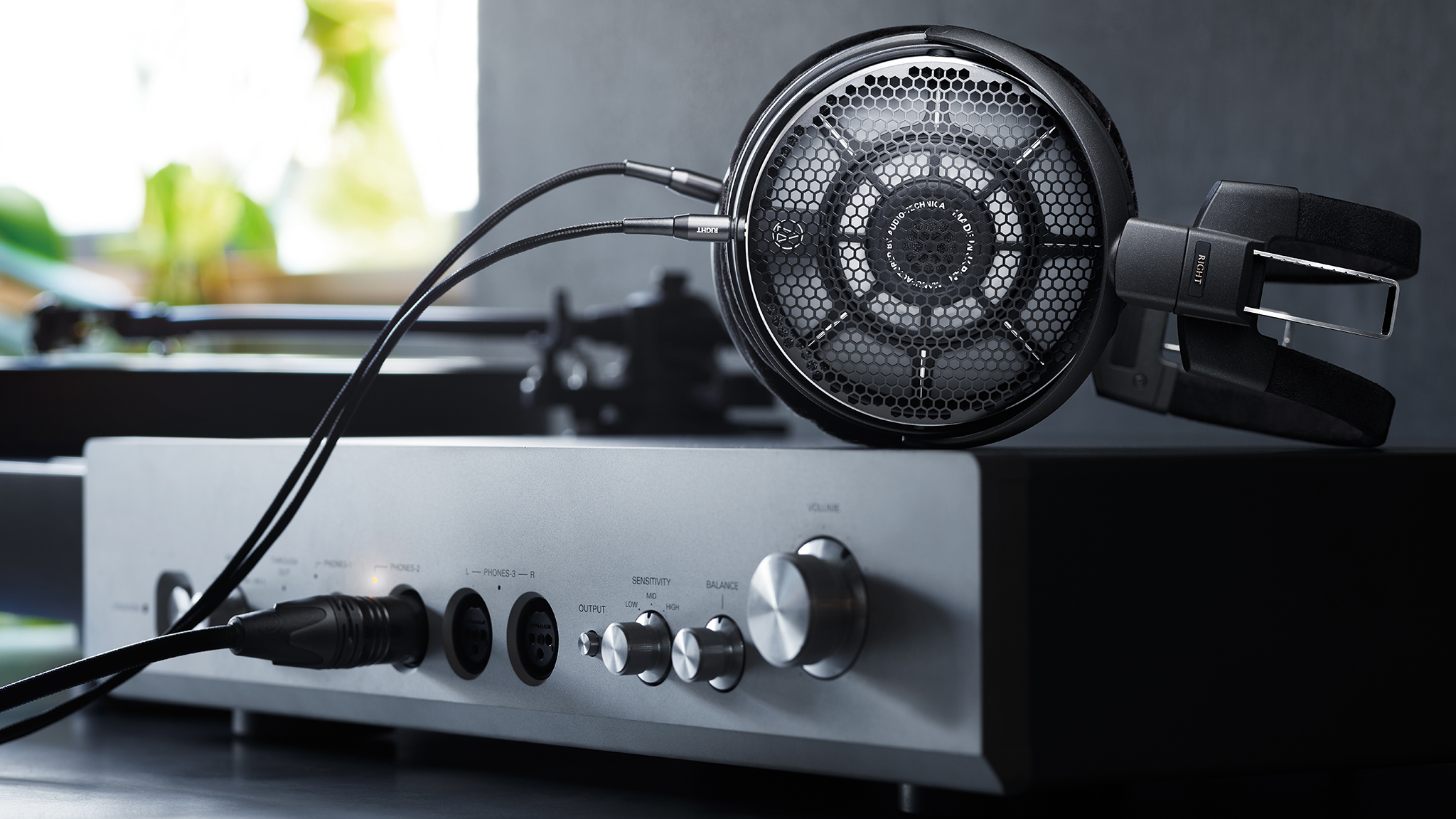 Product photography of the Audio-Technica ATH-ADX7000 over-ear headphones atop a headphone amp with a window in the background