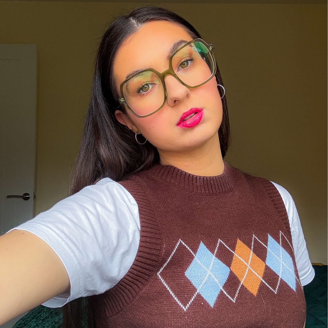 A brown-haired girl with green glasses wearing a brown sweater vest over a white t-shirt poses for the camera
