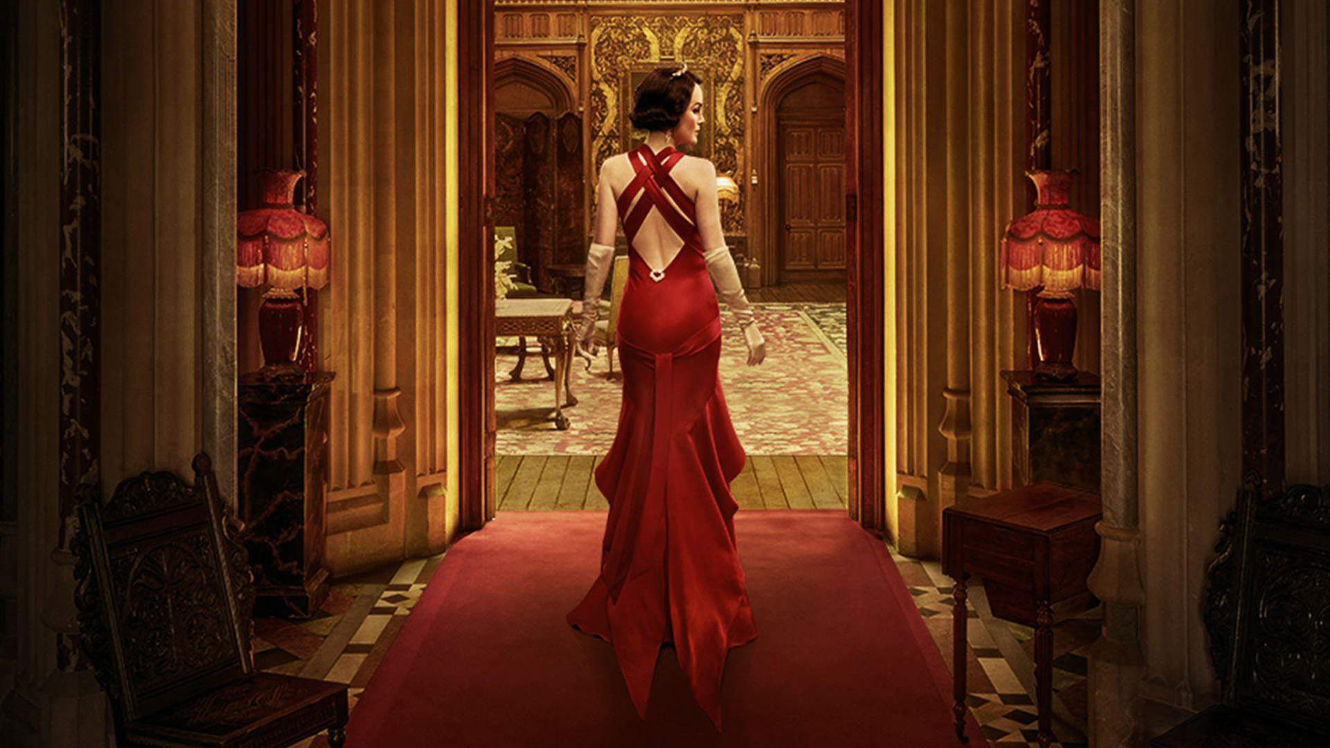 A woman wearing a red dress walks down a red carper in a regal home