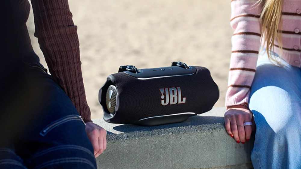 The JBL Xtreme 5 on a wall next to two sitters, and in front of a beach.