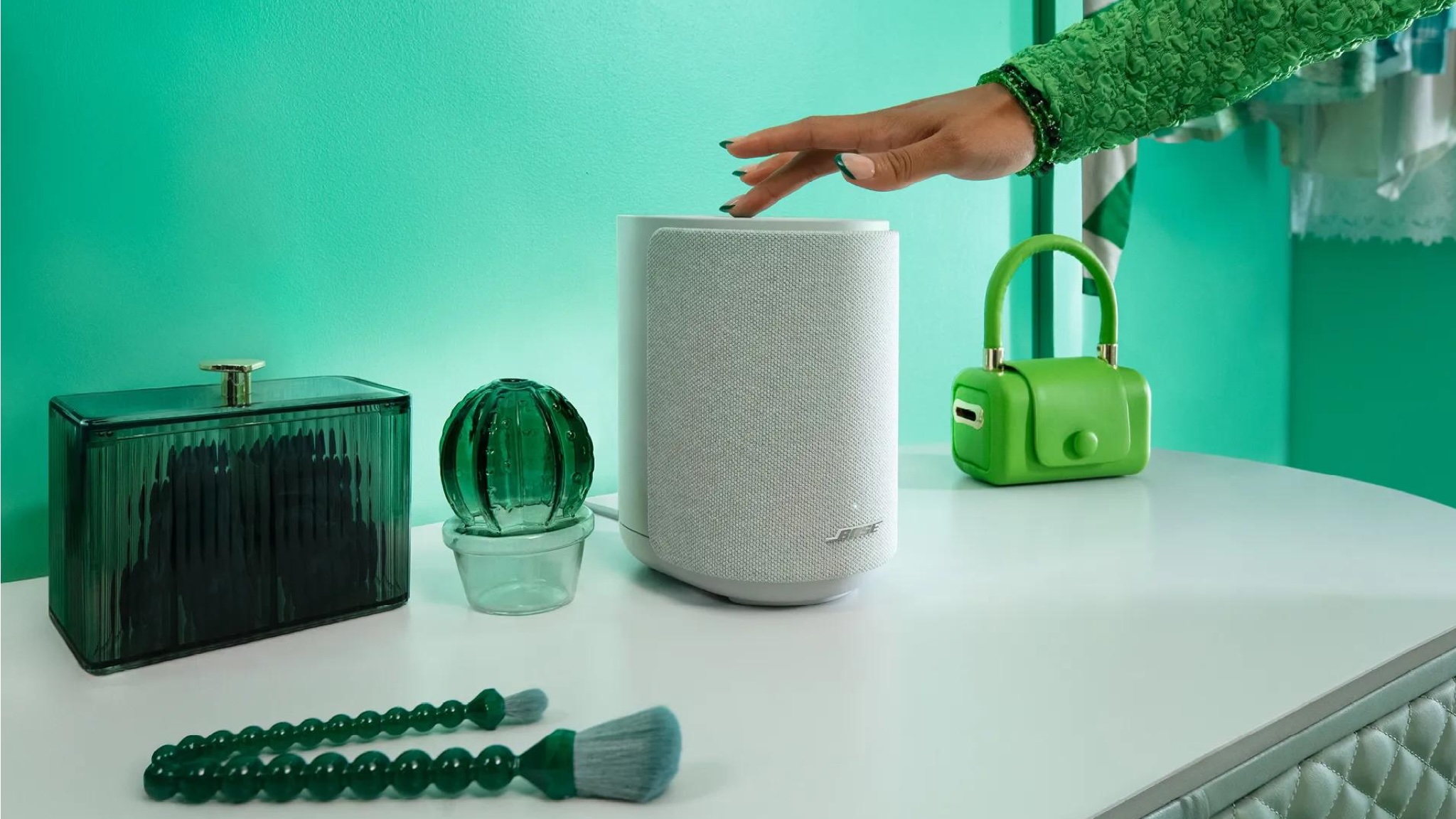 Bose Lifestyle Ultra Speaker on a white surface in front of a green wall. There are green ornaments on the white surface and a person's hand is touching the top of the speaker.