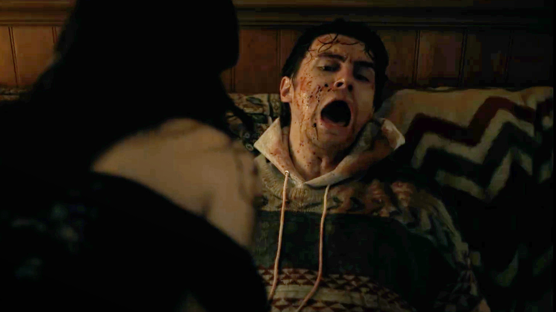 A man screams whilst covered in blood. A woman looms over him, with her back to the camera.